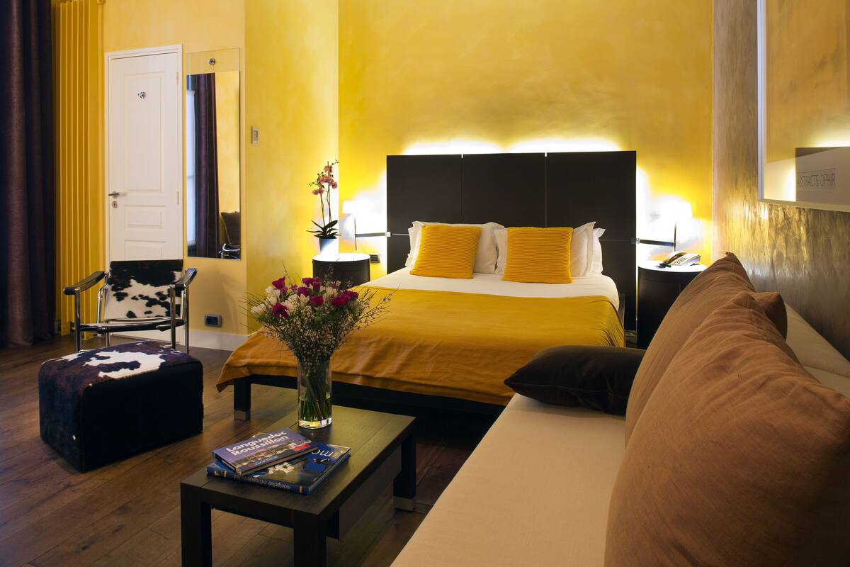 A hotel room with a bed, a sofa, a coffee table, a chair, and a nightstand with a lamp.