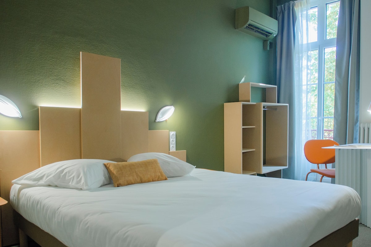 A hotel room with a large bed, a desk with a chair, a wardrobe, and a window with curtains.
