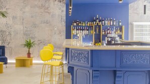 A bar area with a blue accent wall, yellow chairs, and a variety of bottles on shelves. - Hôtel Impérator (Béziers)