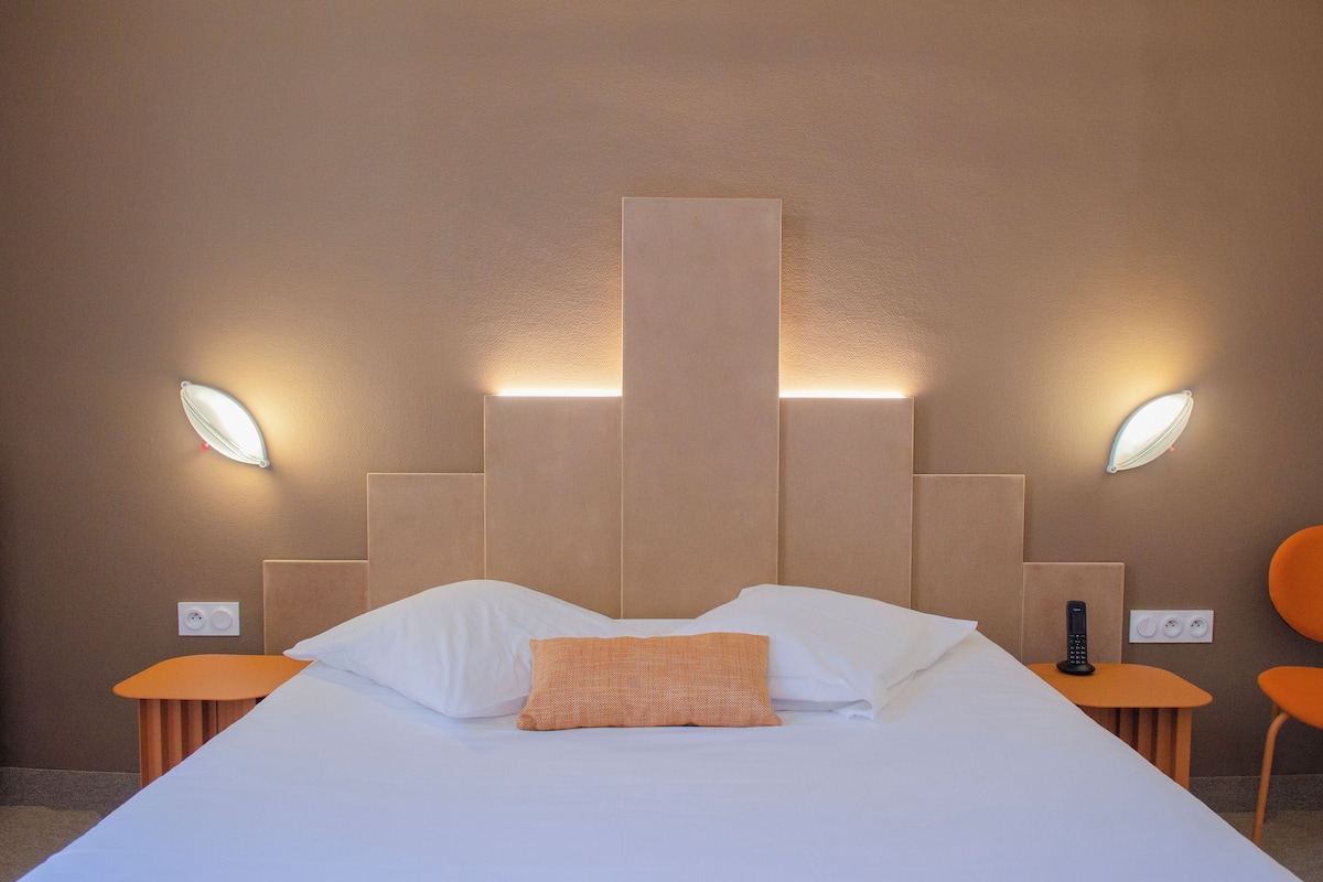 A hotel room with a bed, two bedside tables, a nightstand, and a wall-mounted light fixture.