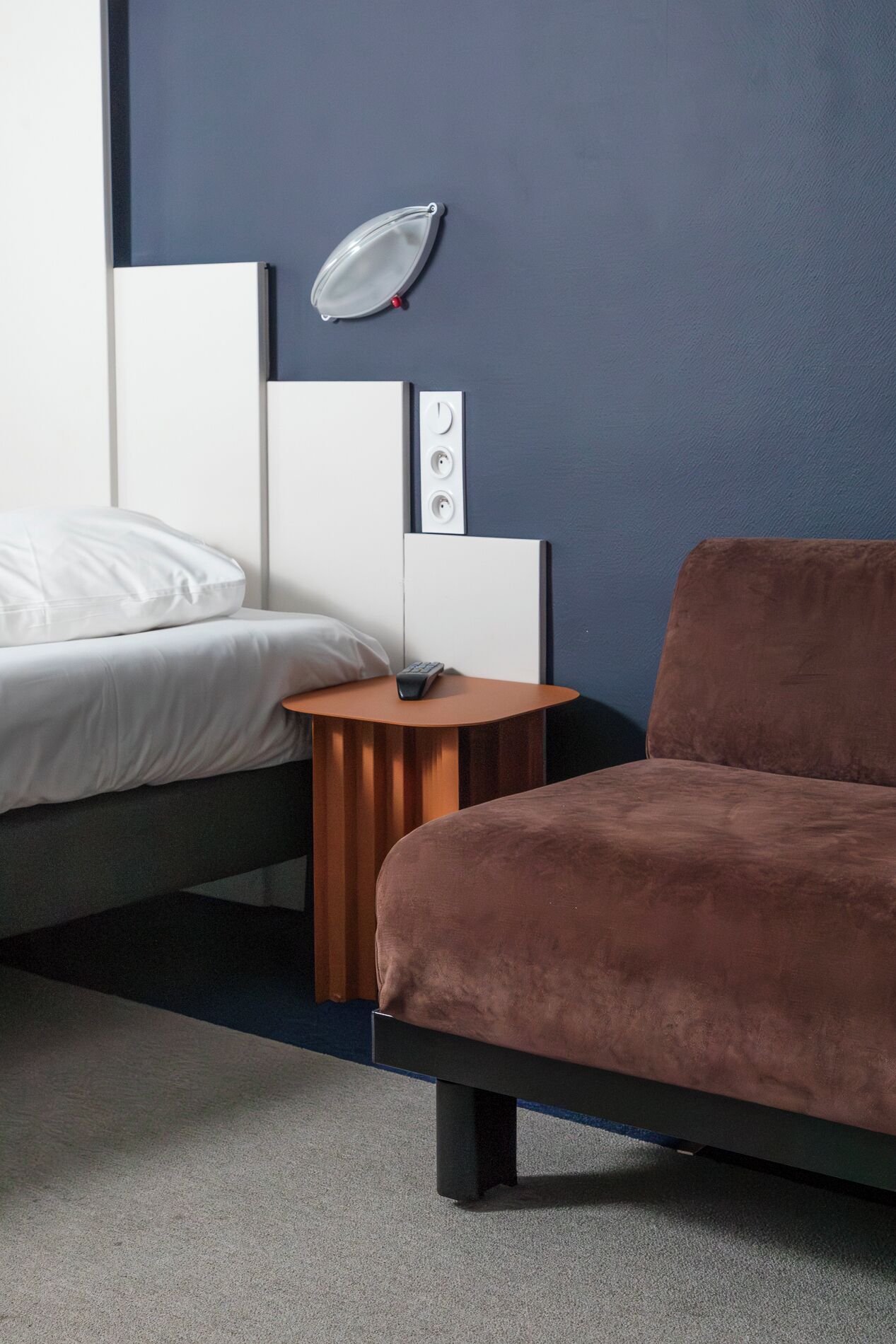 A modern hotel room with a bed, a small wooden side table, a round mirror, and a brown sofa.