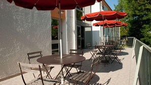 An outdoor patio with tables and chairs, red umbrellas, and a building in the background. - Citotel les Alizes (Eysines)