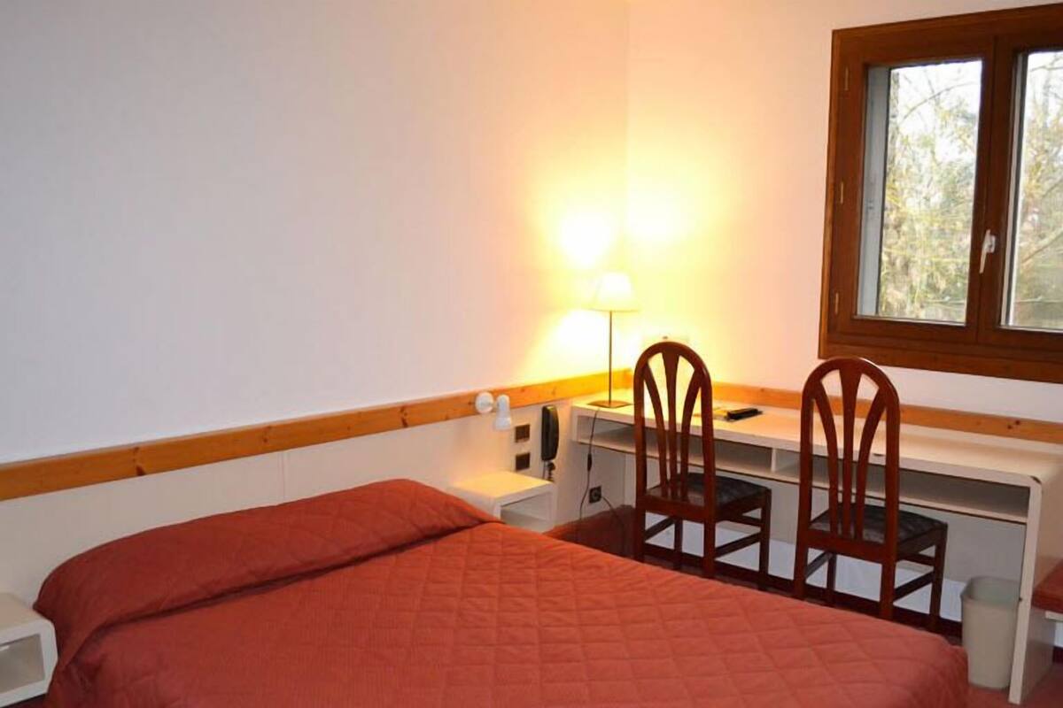 A small, single-bed room with a wooden headboard, a desk with two chairs, a lamp, and a window.