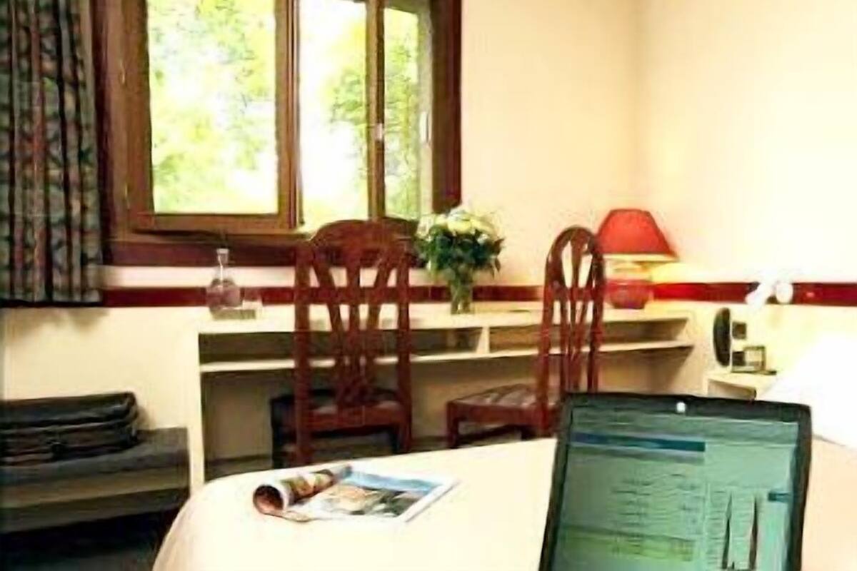 A room with a dining table, chairs, a window with curtains, a vase with flowers, and a computer on the table.