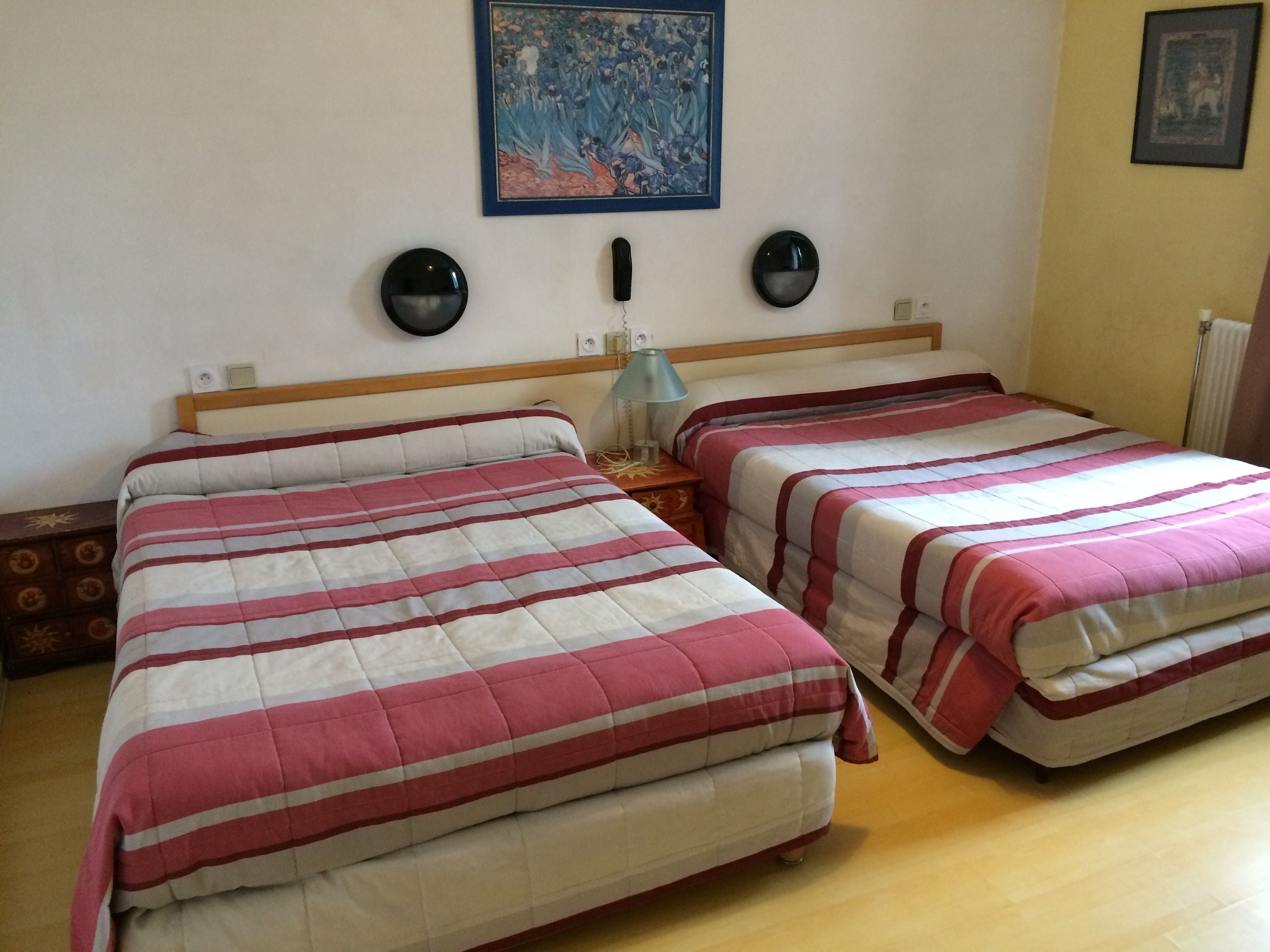 A double room with two beds, each with a striped bedspread, a wooden headboard, bedside tables, and wall-mounted lamps.