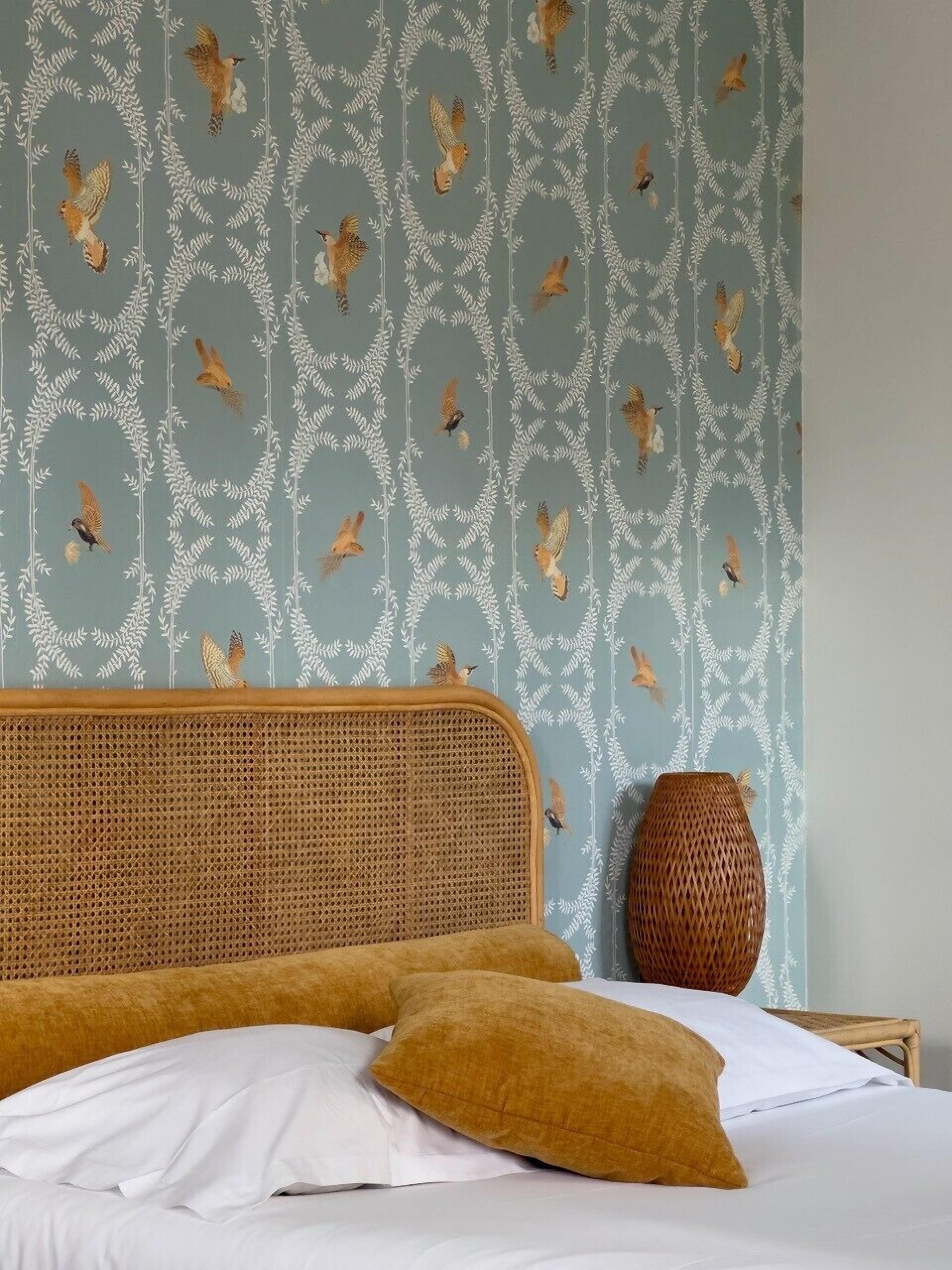 A bed with white and mustard pillows, a wicker headboard, and a wall with a bird pattern.