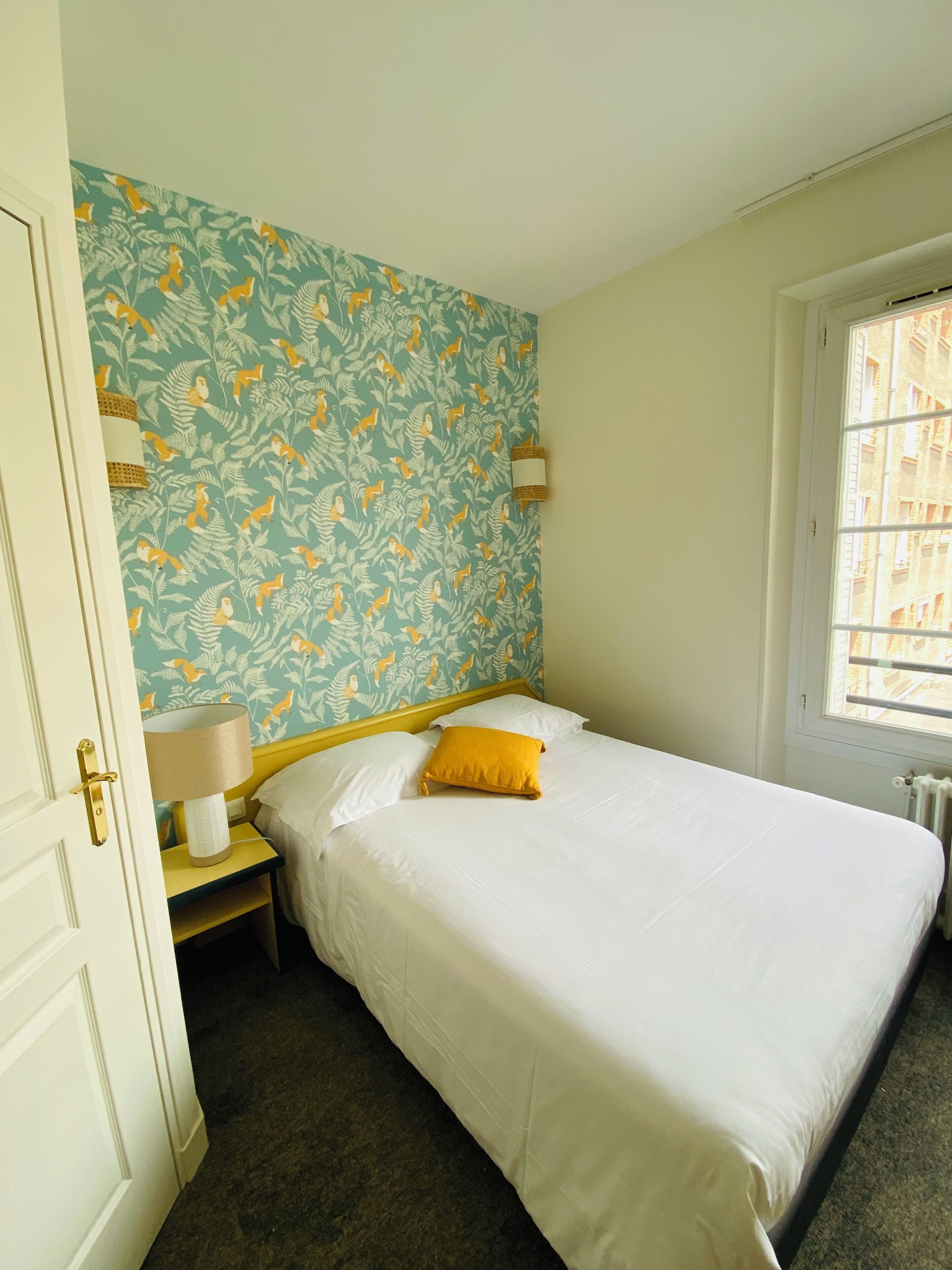 A bedroom with a bed, a nightstand, a lamp, a window, and a patterned wallpaper.
