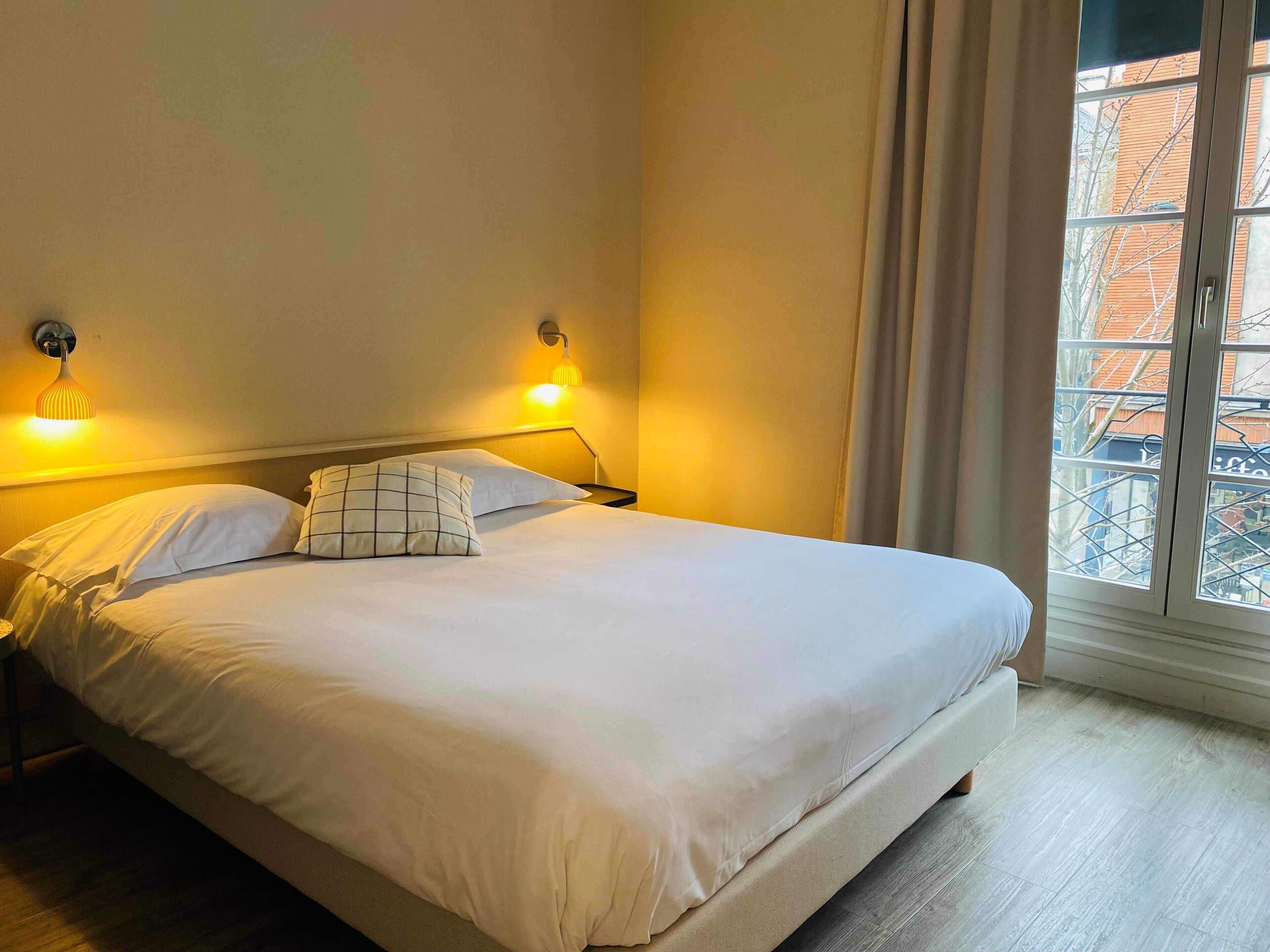 A neatly made bed with white linens and a single decorative pillow, two wall-mounted lamps, and a window with curtains.