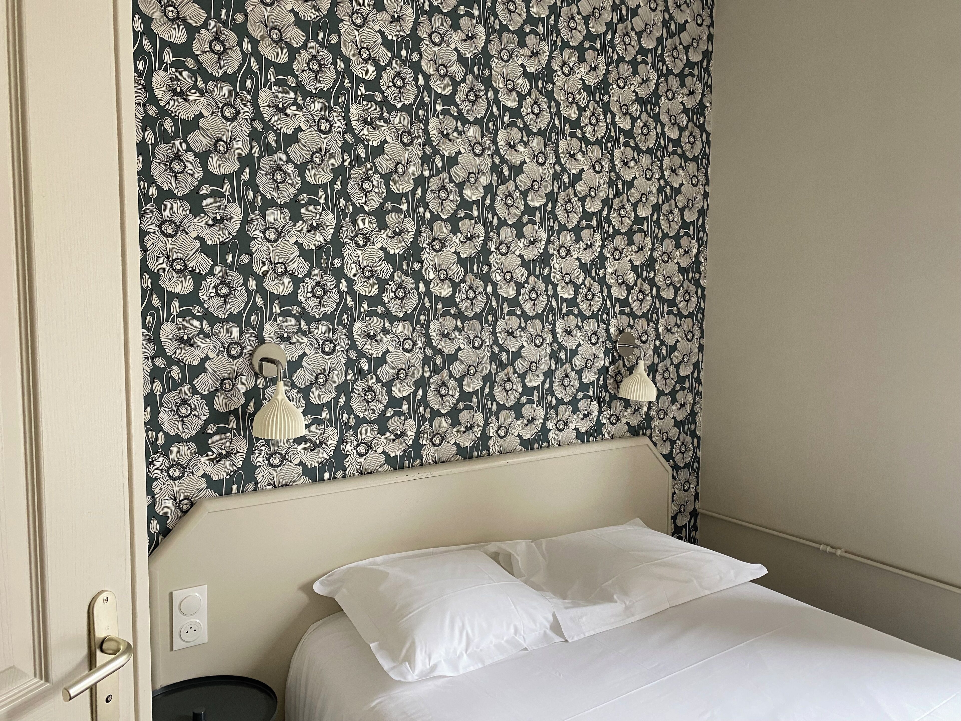 A hotel room with a floral wallpaper, a bed with white bedding, and a wall-mounted lamp.
