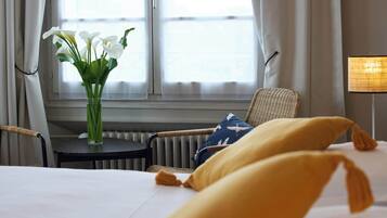 Una habitación de hotel con cama, una silla, una mesita con un florero y una lámpara.