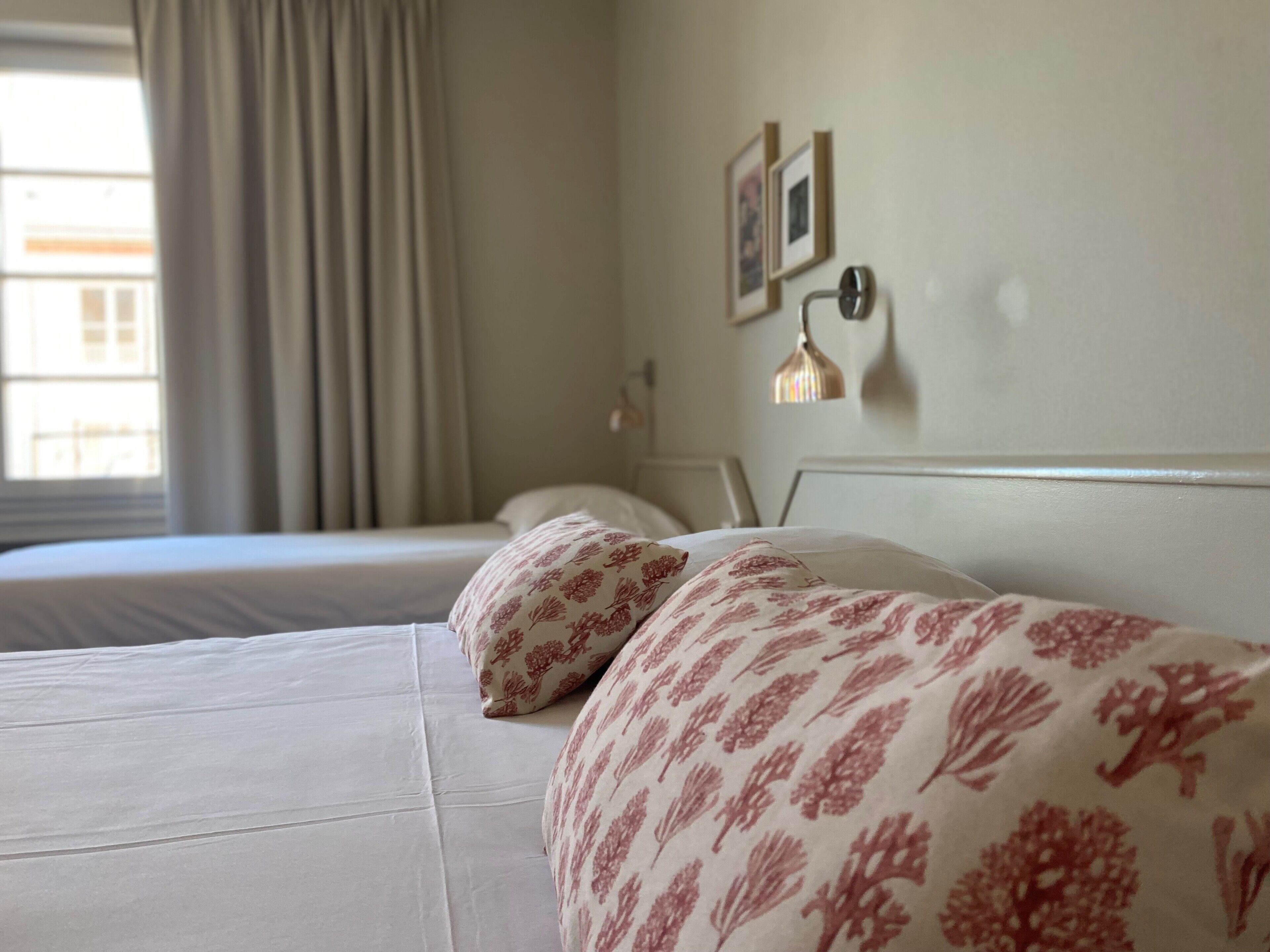 Une chambre d’hôtel avec un lit, une fenêtre avec des rideaux, une lampe fixée au mur et des tableaux encadrés.