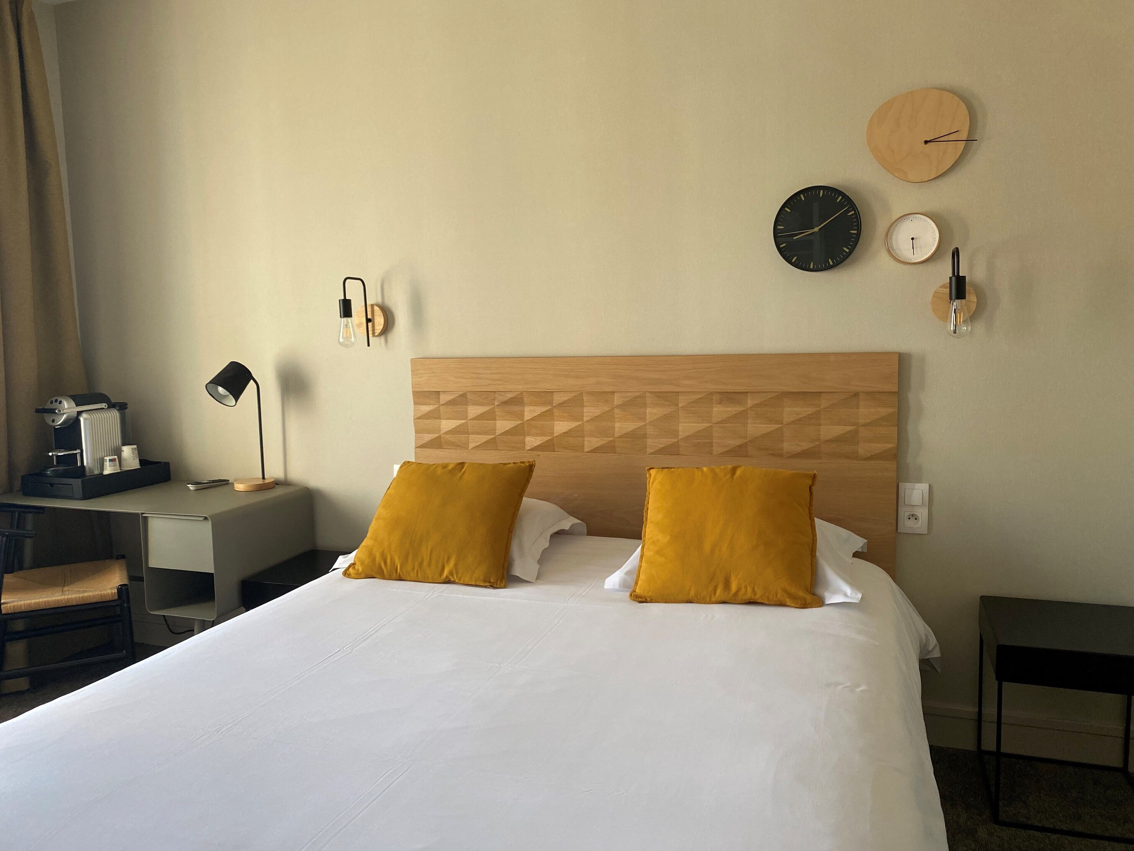 A hotel room with a bed, two mustard pillows, a wooden headboard, a desk with a lamp, a coffee maker, and wall clocks.