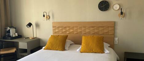 A hotel room with a bed, two mustard pillows, a wooden headboard, a desk with a lamp, a coffee maker, and wall clocks.