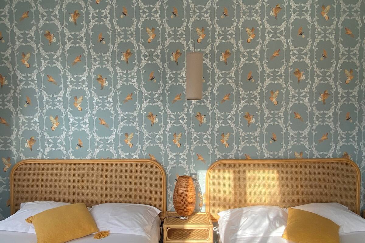 A room with two beds, each with white linens and a single yellow pillow. The walls have a patterned wallpaper with birds and foliage.