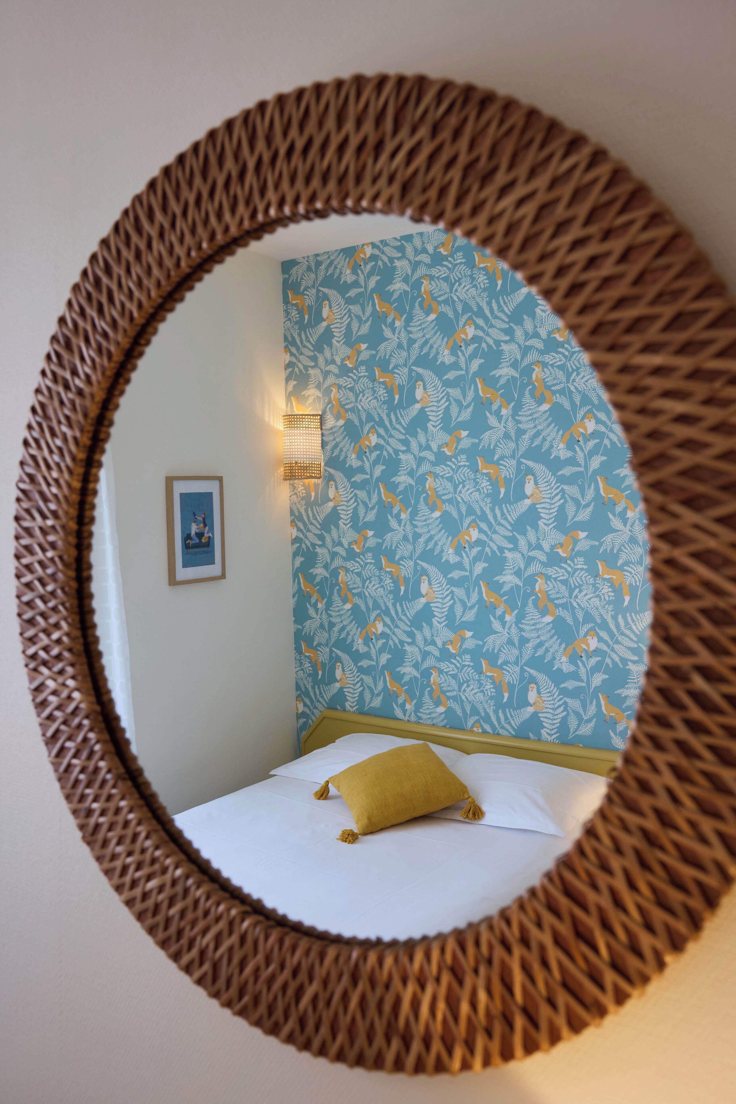 Een bed met witte lakens en een geel kussen, een muur met een blauw bloemenpatroon, en een lijst met een foto aan de muur.