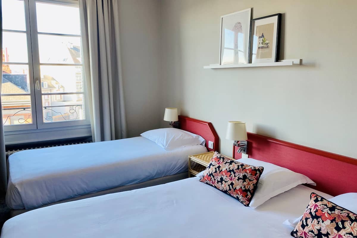 A hotel room with two single beds, a nightstand with a lamp, and a window with curtains.