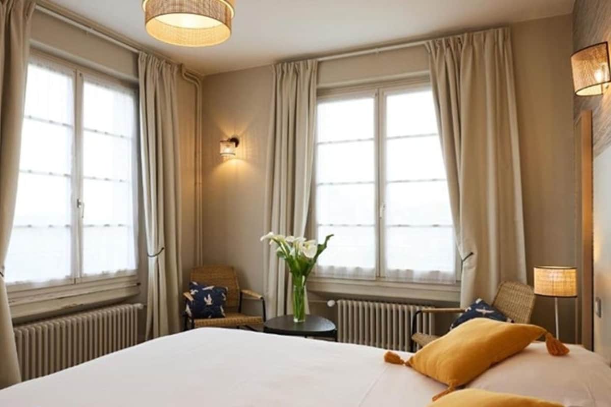 Une chambre d ’hôtel avec un grand lit, deux oreillers, une petite table sur laquelle est posé un vase de fleurs et une lampe.