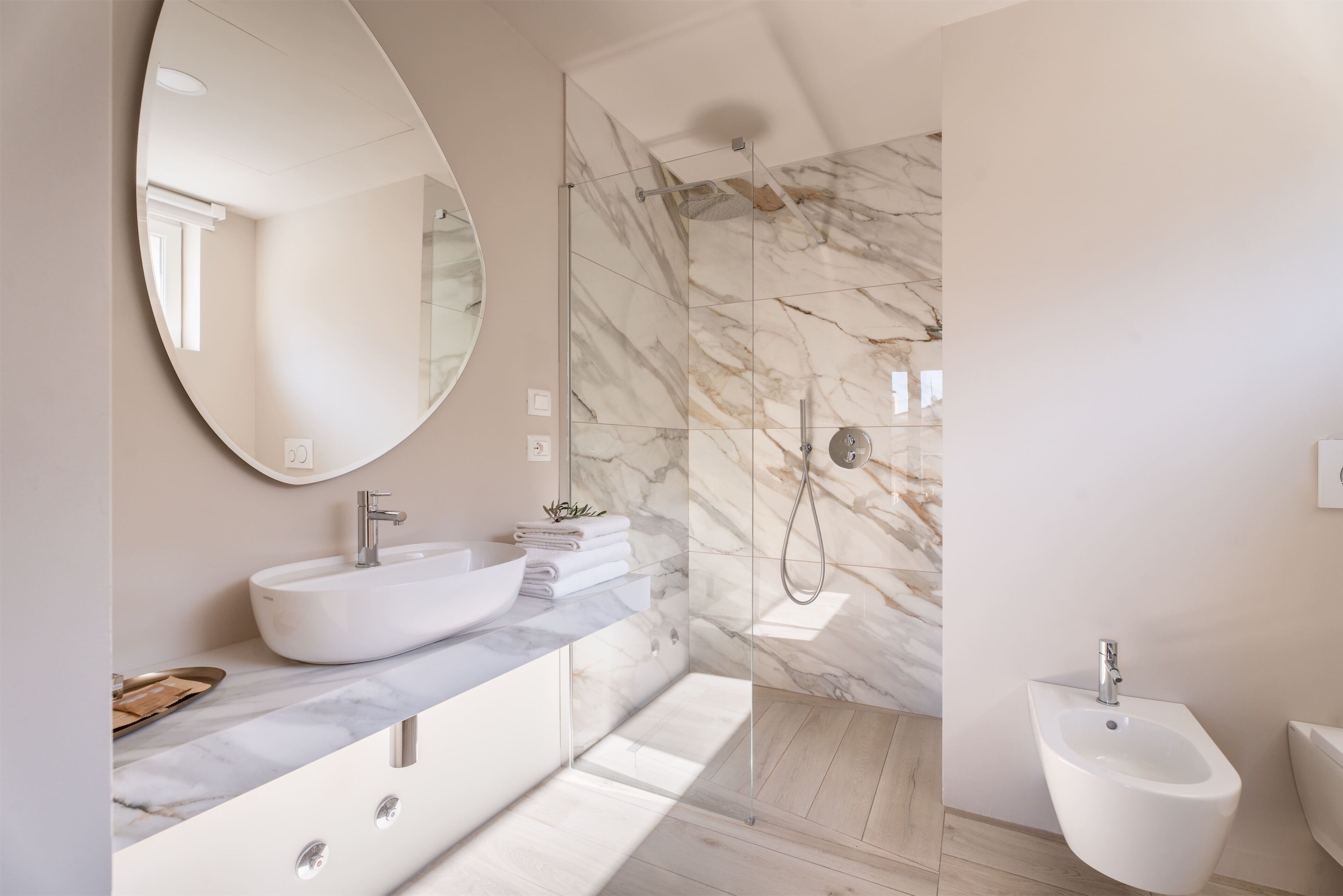 Un baño moderno con un espejo ovalado grande, un lavabo tipo recipiente blanco, un bidé y una ducha con paredes de mármol.
