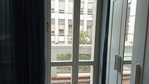 A view through a window with a metal handle, a balcony with a railing, and a building with multiple windows in the background. - New Paradise (Rome)