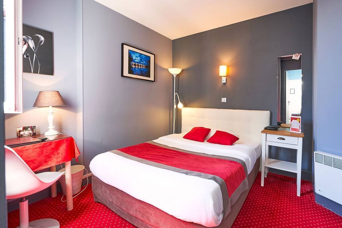 A hotel room with a bed, a desk, a chair, a nightstand, a lamp, a framed picture, and a mirror.