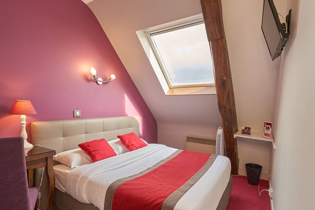 A bedroom with a bed, a nightstand, a lamp, a window, and a shelf with a picture frame.