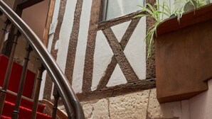 Una escalera con peldaños alfombrados en rojo conduce a una plataforma. Una barandilla de madera se encuentra a la izquierda, y una planta en maceta a la derecha. La pared presenta piedra vista y entramado de madera.