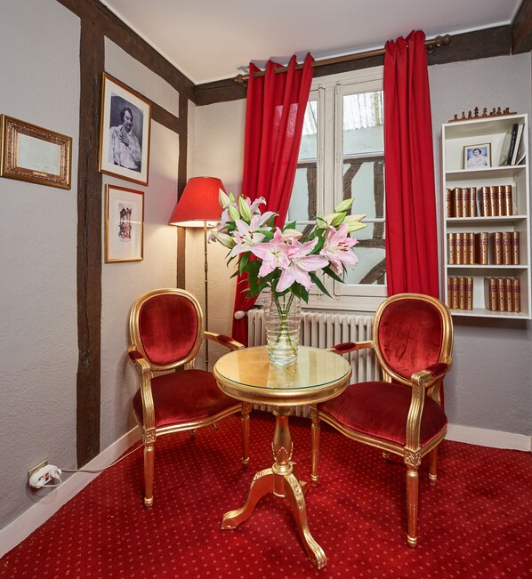 Une pièce avec une table ronde, deux chaises, un vase de fleurs, une lampe, une bibliothèque et un tableau encadré au mur.