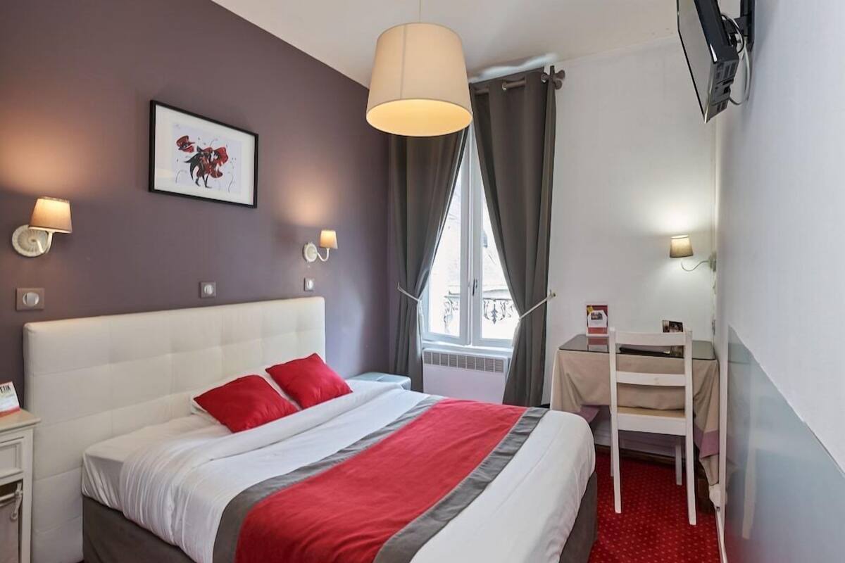 A hotel room with a large bed, a desk, a chair, two wall-mounted lamps, a window with curtains, and a painting on the wall.