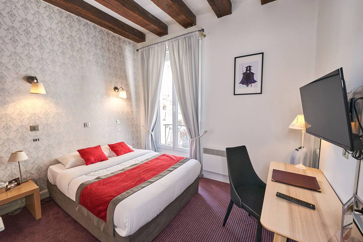 A hotel room with a bed, a desk with a laptop, a chair, a TV, and a window with curtains.