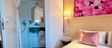 A hotel room with a shower, sink, toilet, and a bed with a large floral wall decoration.