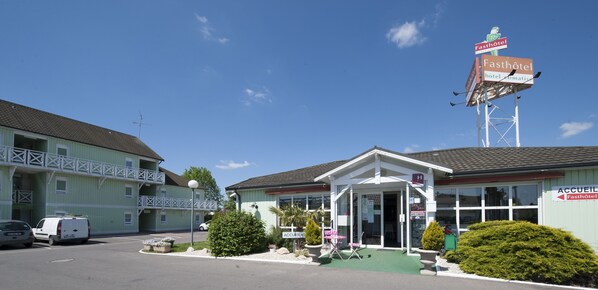 A two-story building with a sign for 'Fasthotel' and 'ACCUEIL'. There is a covered entrance with a glass door, a small outdoor seating area, and a parking lot with a white van and a car.