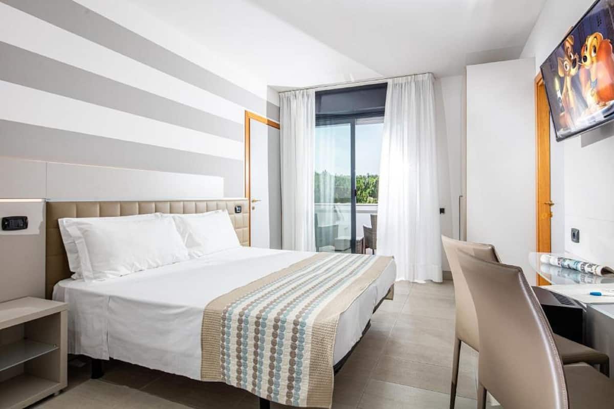 A hotel room with a bed, a desk, and a chair. There is a window with curtains, a television mounted on the wall, and a view of greenery outside.