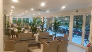 A spacious indoor area with wicker furniture, large windows, and a view of the outdoors. - Hotel Helvetia (Lignano Sabbiadoro)