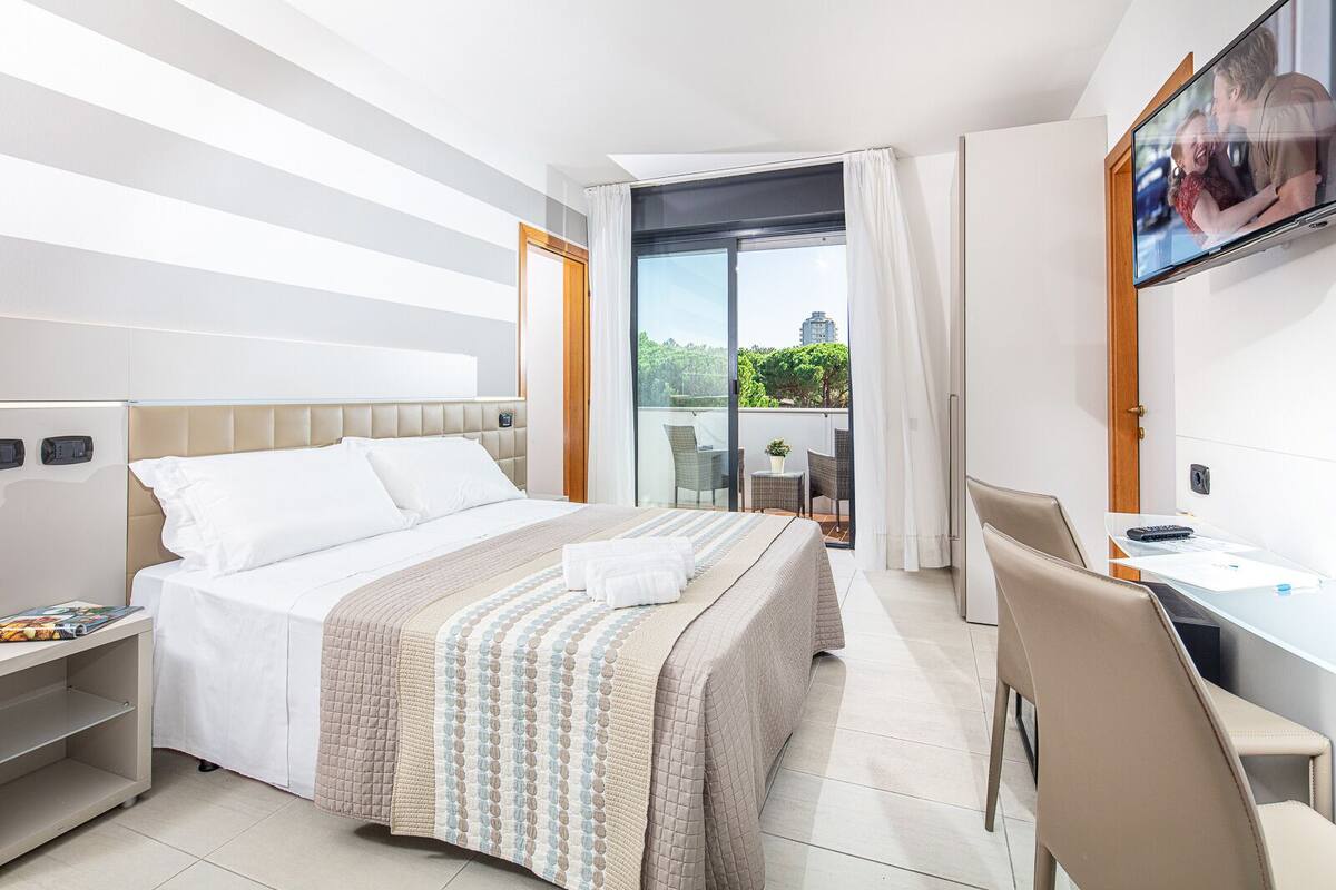 A modern hotel room with a large bed, a desk, and a balcony with a city view.