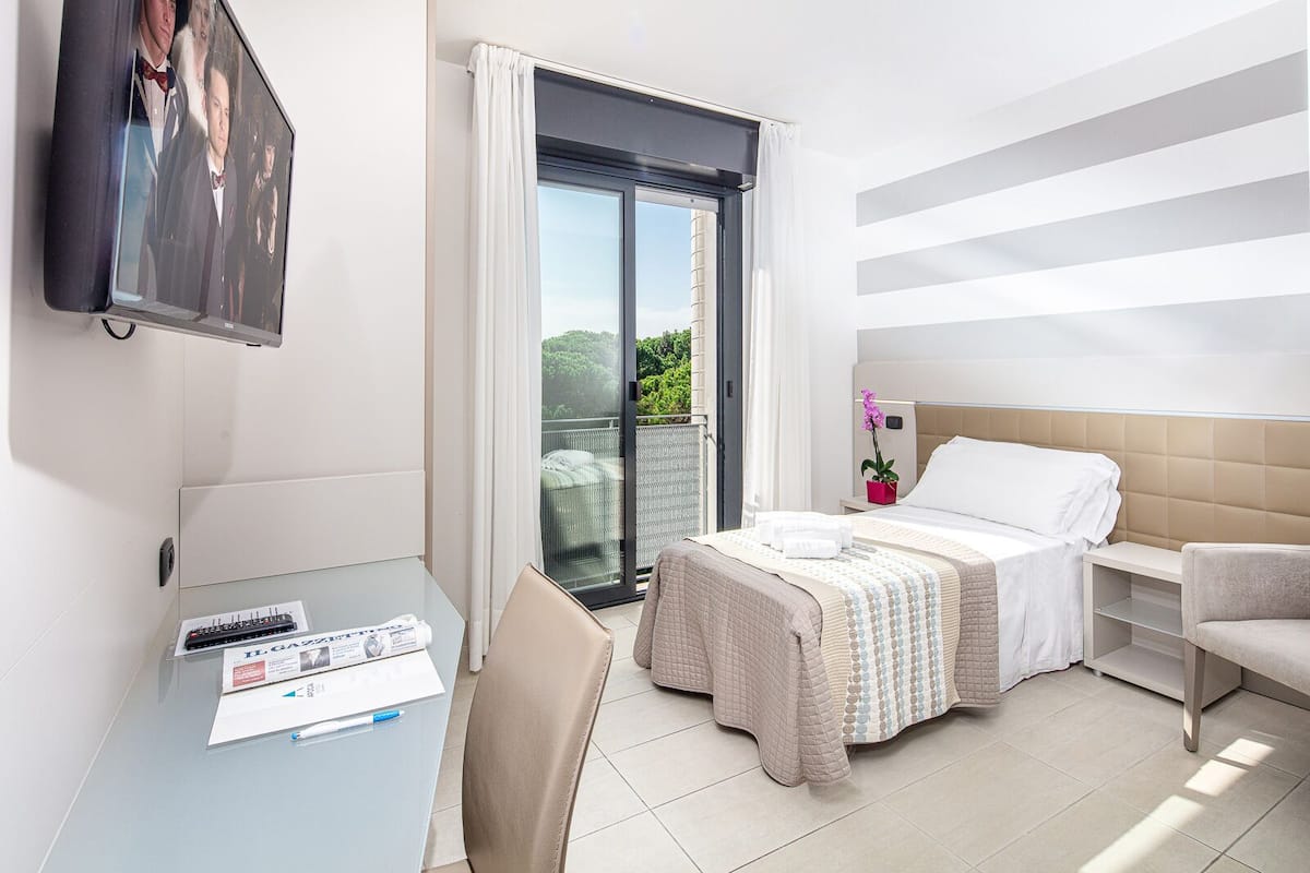 A hotel room with a bed, a desk with a chair, a TV, and a balcony with a view of greenery.