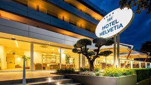 A modern hotel entrance with a sign reading "HH HOTEL HELVETIA," steps leading up to the entrance, and a well-lit outdoor area with a tree and plants. - Hotel Helvetia (Lignano Sabbiadoro)