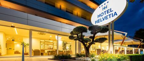 Una entrada de hotel moderna con un cartel que dice “HH HOTEL HELVETIA”, escalones que conducen a la entrada y un área exterior bien iluminada con un árbol y plantas.