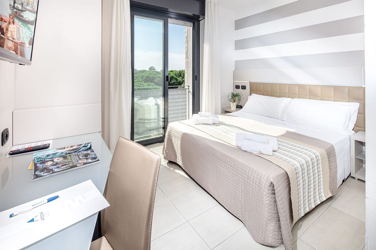 A hotel room with a bed, a desk with a chair, a window with a view of greenery, and a sliding door.
