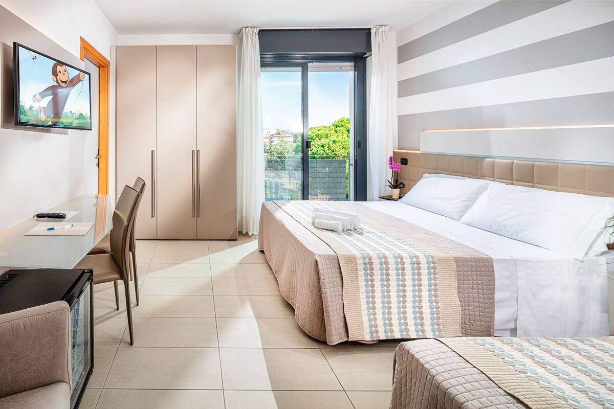 A hotel room with a bed, a desk, a chair, a TV, and a view of greenery outside.