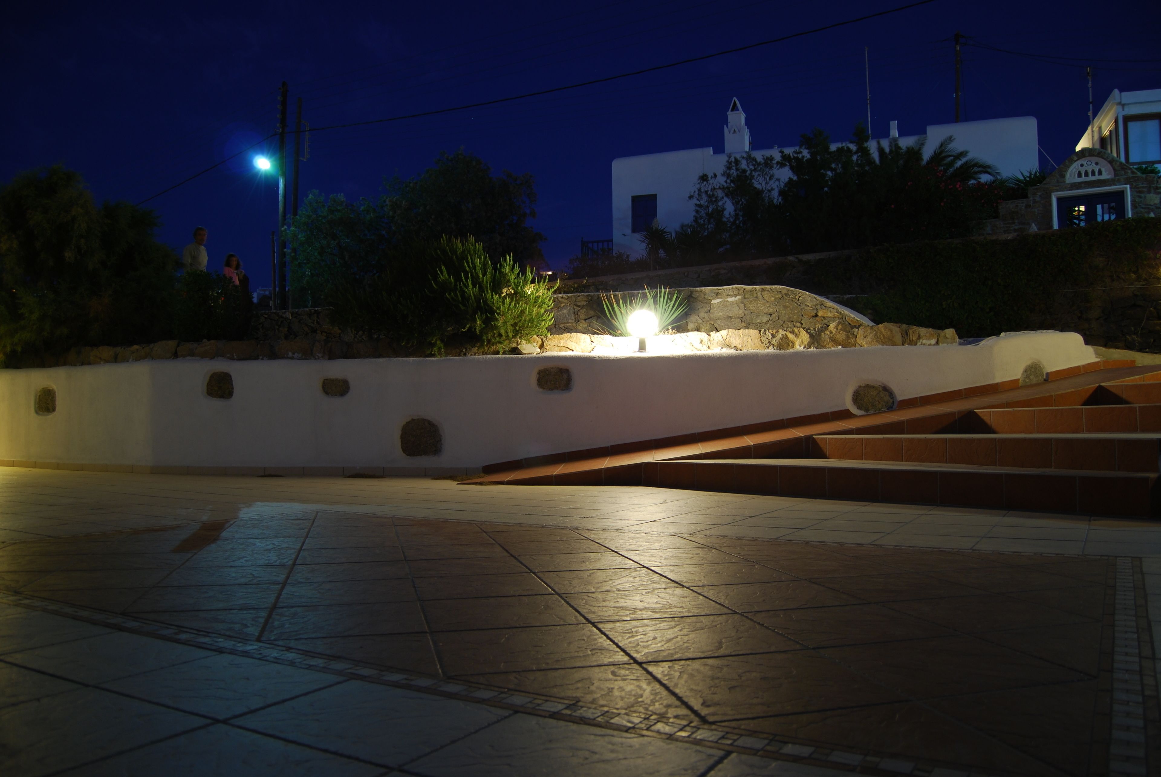 Vista nocturna de una plaza con un muro de piedra, un farol y un edificio con fachada blanca al fondo.