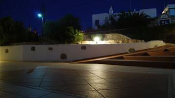 Vista nocturna de una plaza con un muro de piedra, un farol y un edificio con fachada blanca al fondo.