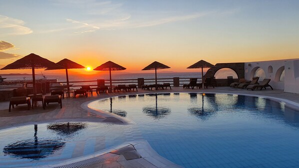 Zona junto a la piscina con sillones y sombrillas, con vista al mar al atardecer.