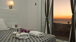 A hotel room with a bed, a small table with a lamp, a window with curtains, and a view of the sunset. - Gorgona Hotel (Mykonos)