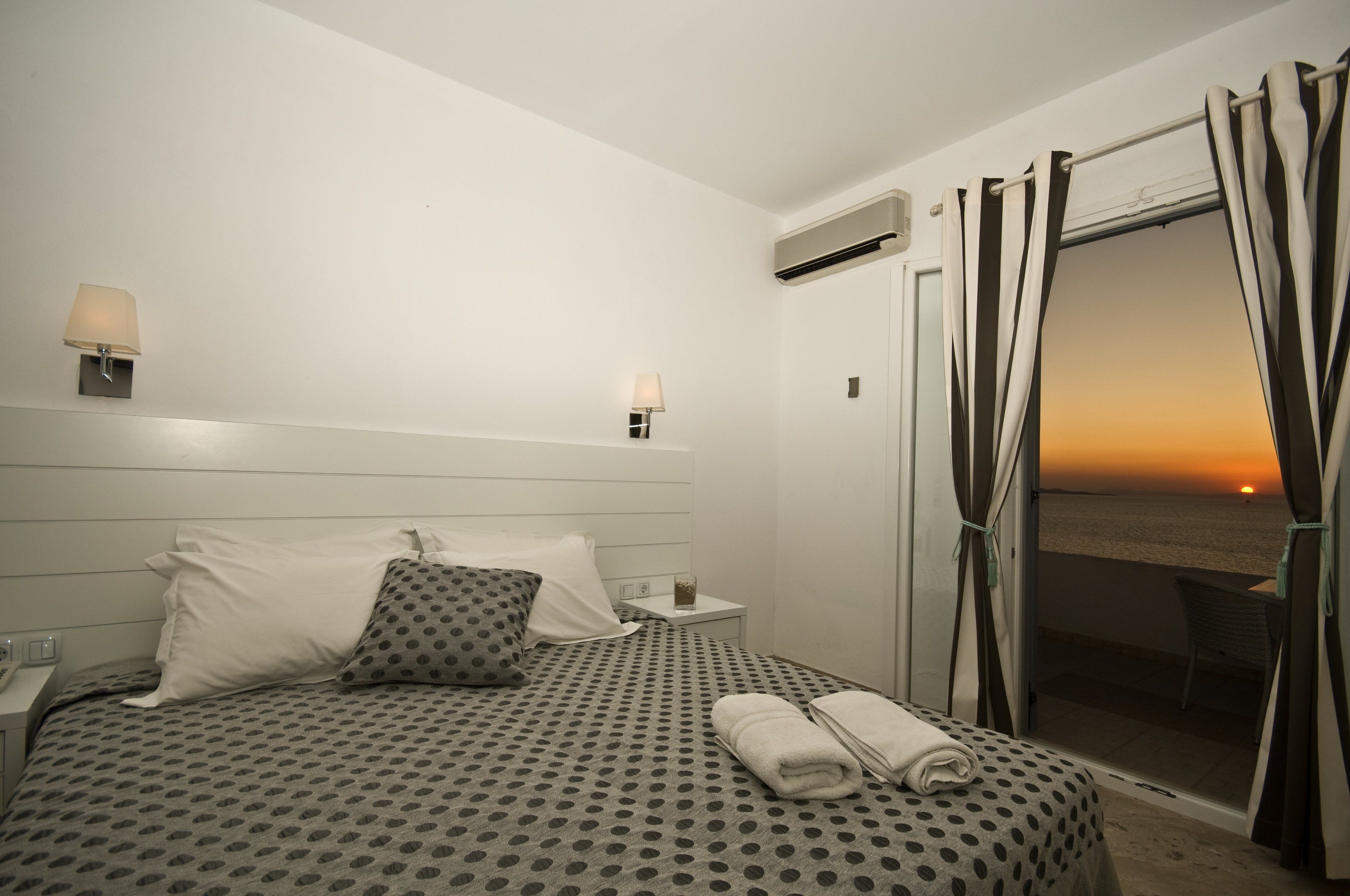 Una camera d'albergo con un letto, un comodino, una lampada e vista sul tramonto sull'oceano.