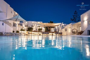 Una piscina con tumbonas y sombrillas, rodeada de edificios blancos con balcones y zonas de estar al aire libre.
