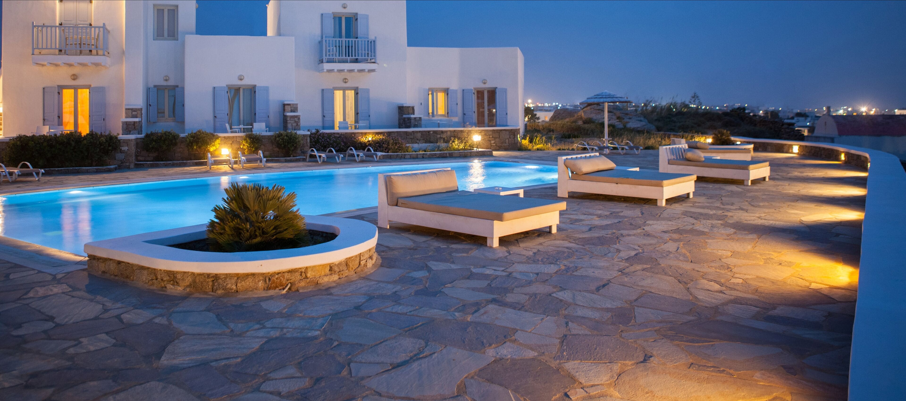 A pool area with lounge chairs and a stone floor, surrounded by a white building with balconies.