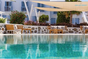 Un área junto a la piscina con sillones y sombrillas, un muro de piedra y un edificio con contraventanas azules al fondo.