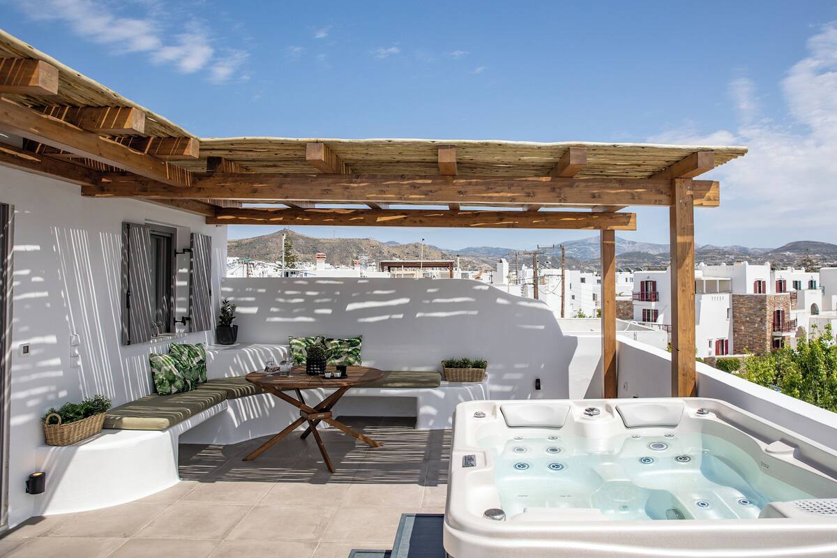 A rooftop terrace with a wooden pergola, a seating area with cushions, a hot tub, and a view of the cityscape.