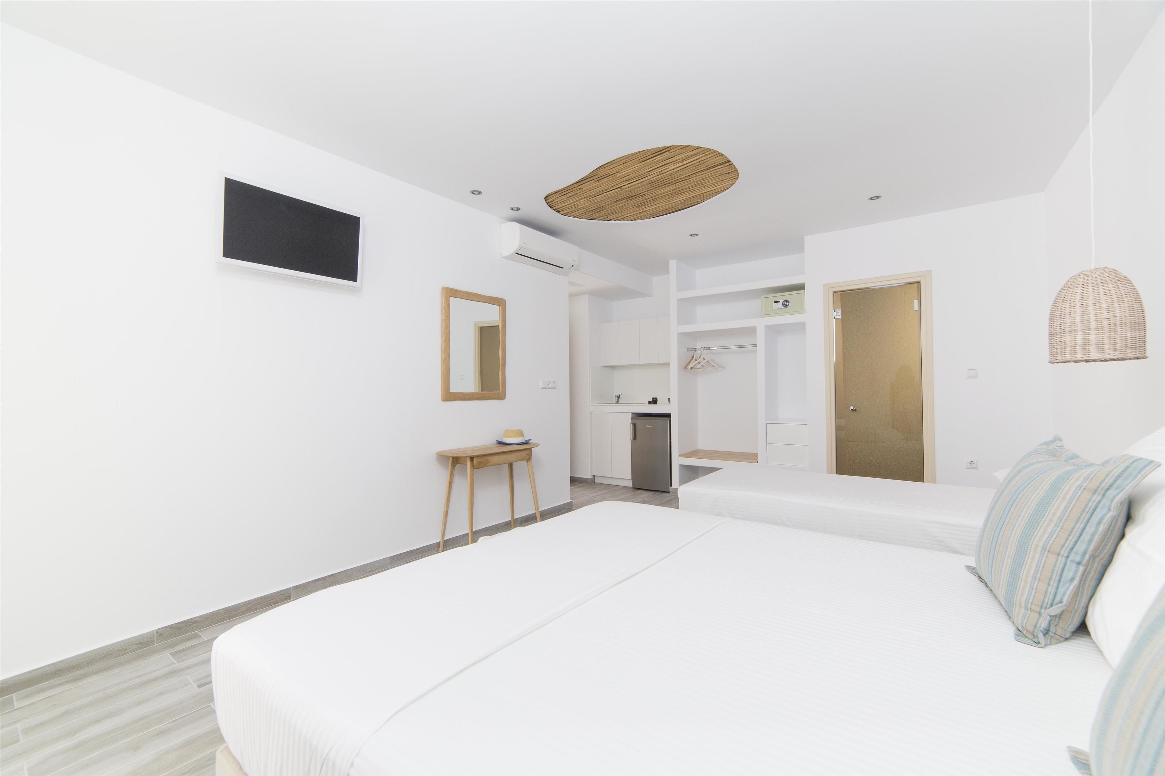 Une chambre moderne avec un grand lit, une petite table, une armoire et un coin cuisine à l’arrière-plan.