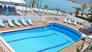 A swimming pool with blue water, surrounded by lounge chairs and a view of the sea and mountains.